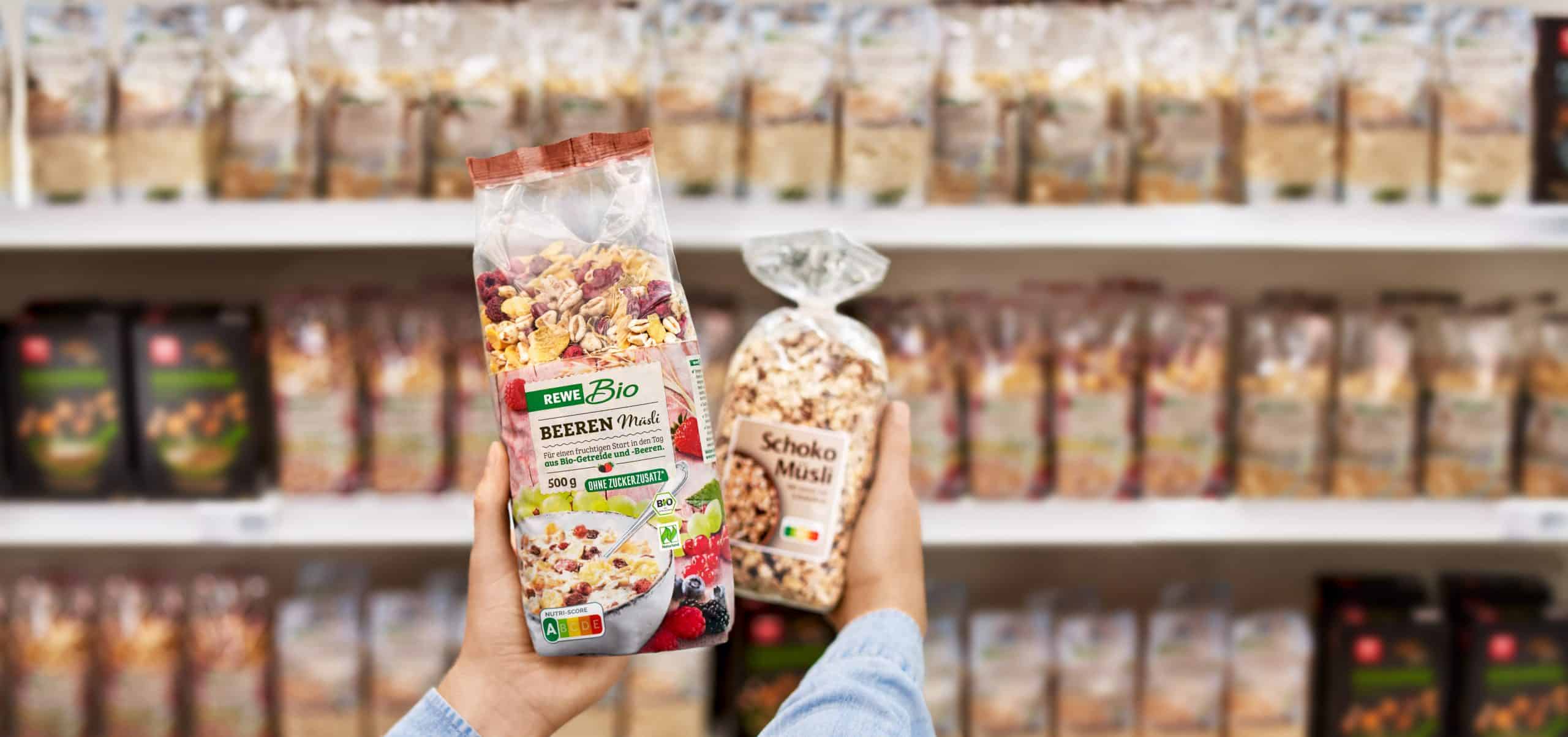 Zwei verschiedene Müslis werden vor einem Supermarkt-Regal hochgehalten. Bei dem vorderen Müsli ist der Nutri-Score zu sehen.
