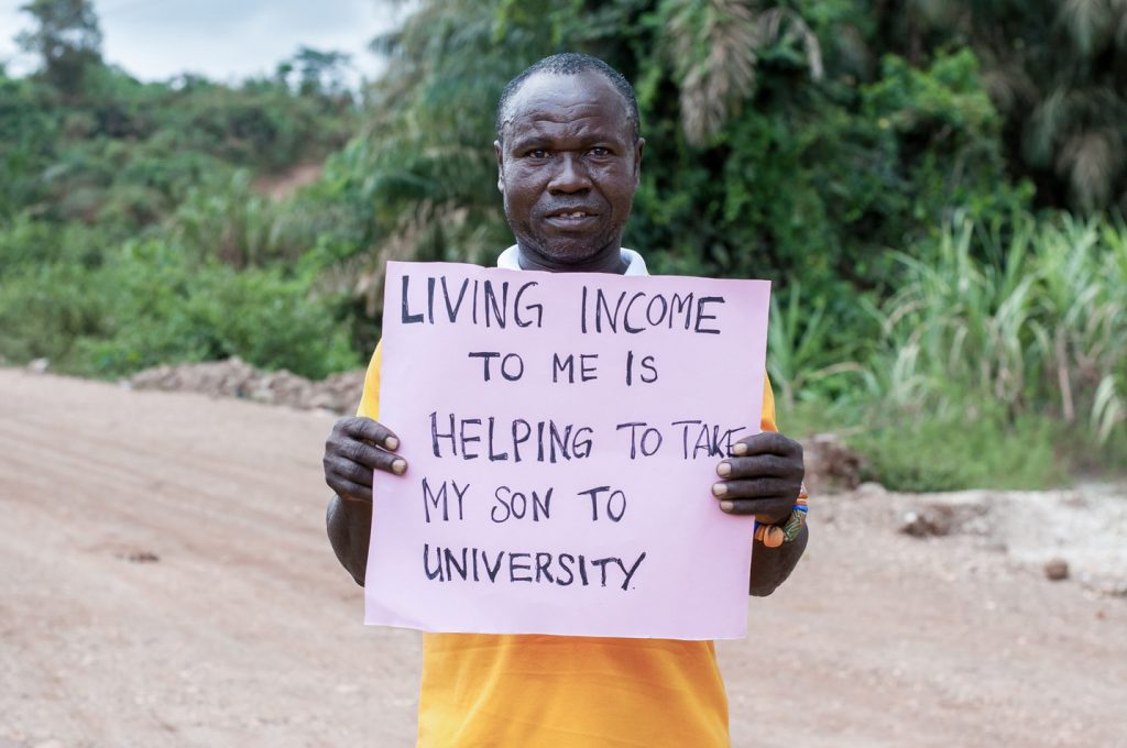 Ein Farmer hält ein Schild hoch, auf dem geschrieben steht: „Existenzsichernder Lohn bedeutet für mich, meinen Sohn dabei zu unterstützen, die Universität besuchen zu können.“