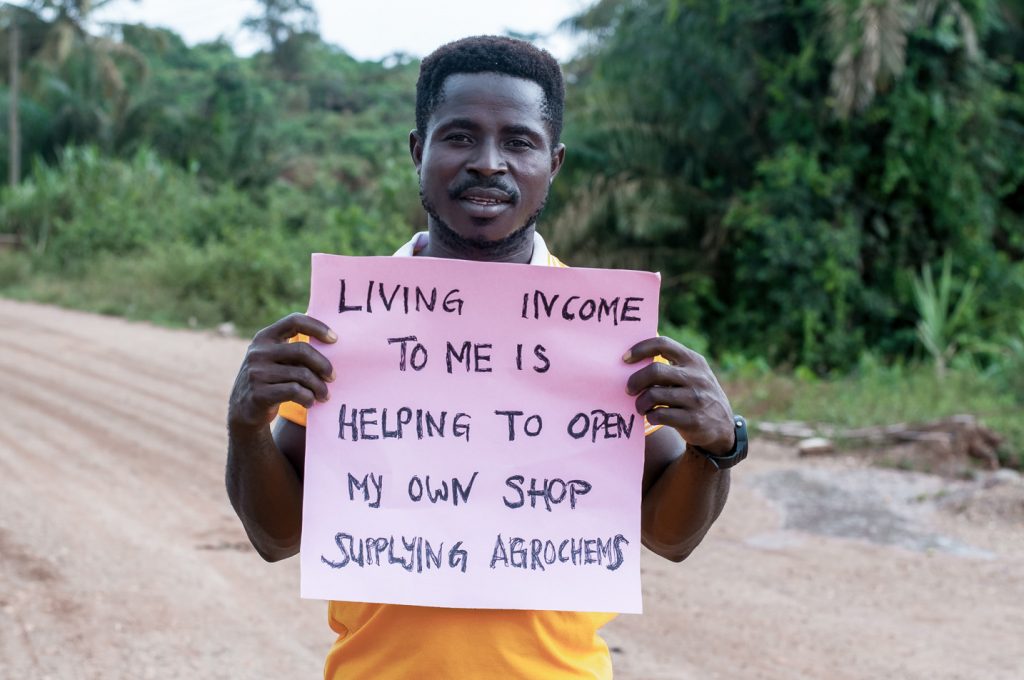 Ein Farmer hält ein Schild hoch, auf dem geschrieben steht: „Existenzsichernder Lohn bedeutet für mich, mein eigenes Geschäft mit Agrochemie eröffnen zu können.“