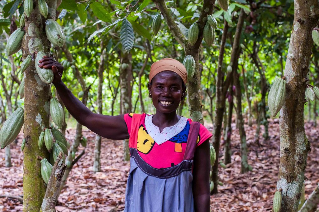 A cacao farmer stands next to a cacao plant and puts her hand on a cacao bean.