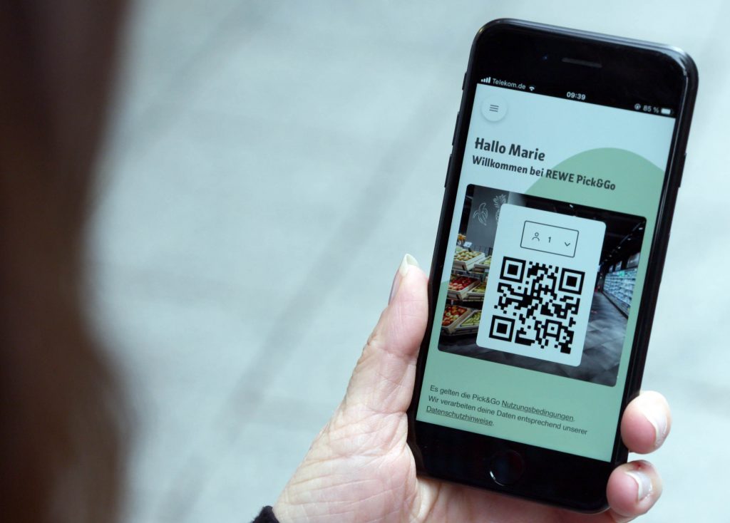 Eine Frau hält ein Handy in die Kamera, darauf ist ein QR Code zu sehen und "Hallo Marie, Willkommen bei REWE Pick & Go" zu lesen