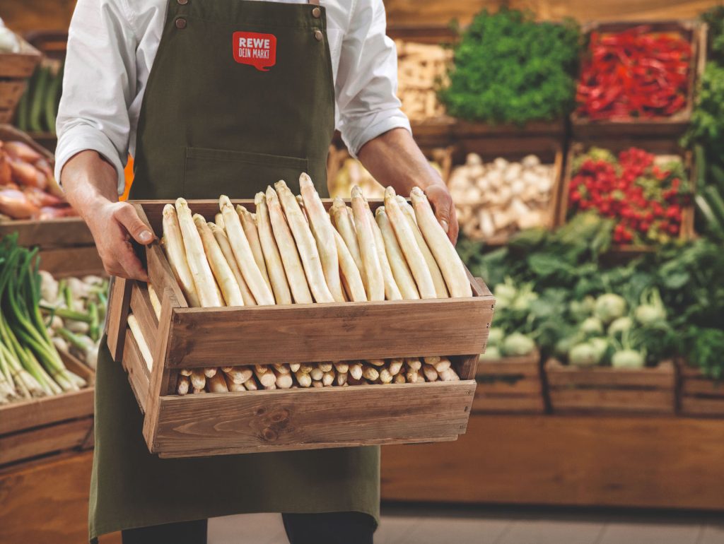 Eine Holzkiste mit Spargel in einem REWE Markt. In den Regalen im Hintergrund liegt unverpacktes Gemüse