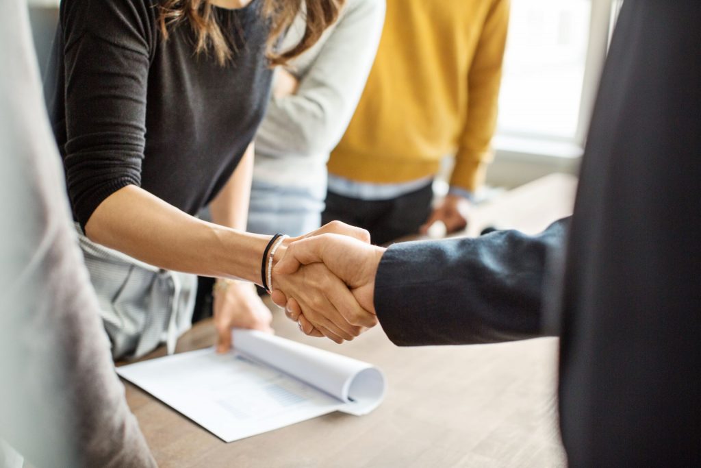 Ein Handschlag: Zwei Personen schütteln sich die Hände, die eine Person hält einen Vertrag in der Hand.