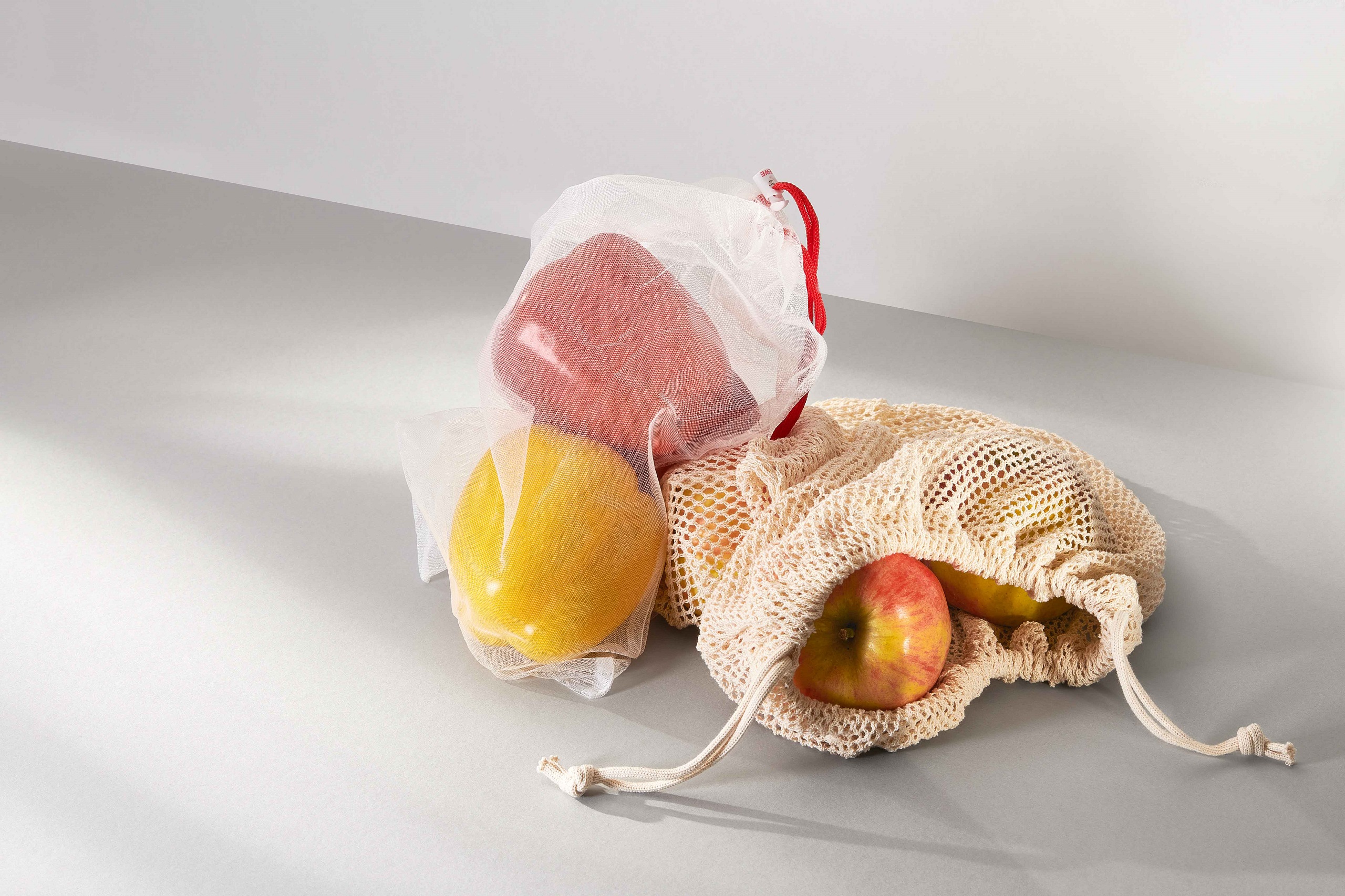 Two reusable fresh nets on a white surface. One with peppers and one with apples.