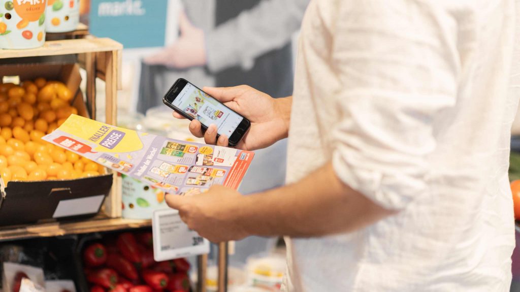 Person hält gedruckten Handzettel und digitalen Handzettel im Supermarkt nebeneinander.