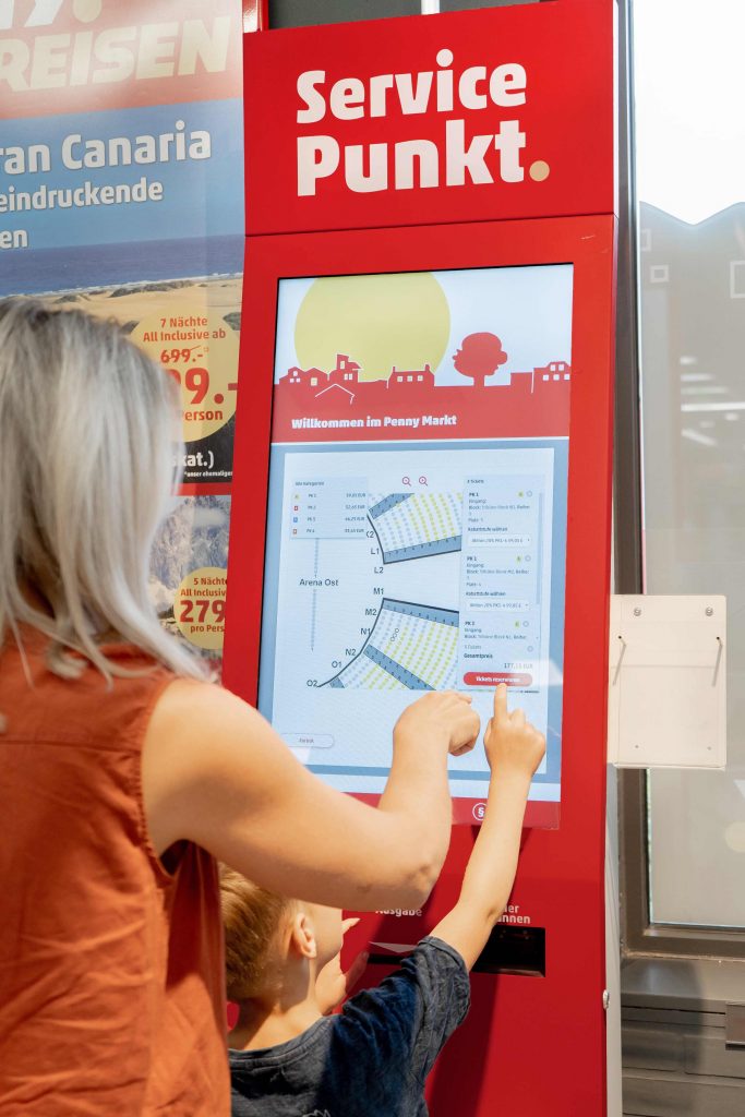 Frau und Kind von hinten, welche an einem Servicepunkt Automaten in einem Penny Markt Tickets für eine Veranstaltung auswählen.