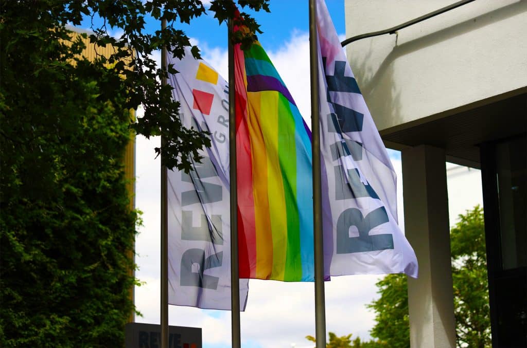 Eine Regenbogenflagge und zwei REWE Group Flaggen, die im Wind wehen