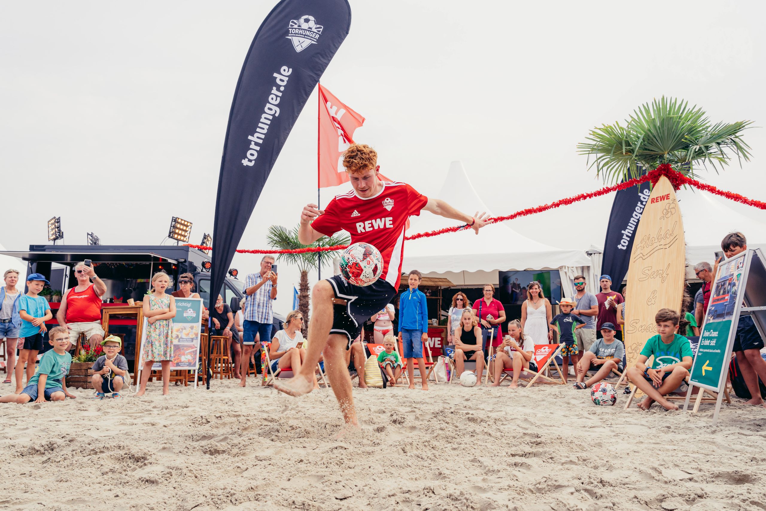 Fussballspieler mit REWE Trikot an einem Strand mit der Torhunger-Aktionsflagge vor einem Publikum