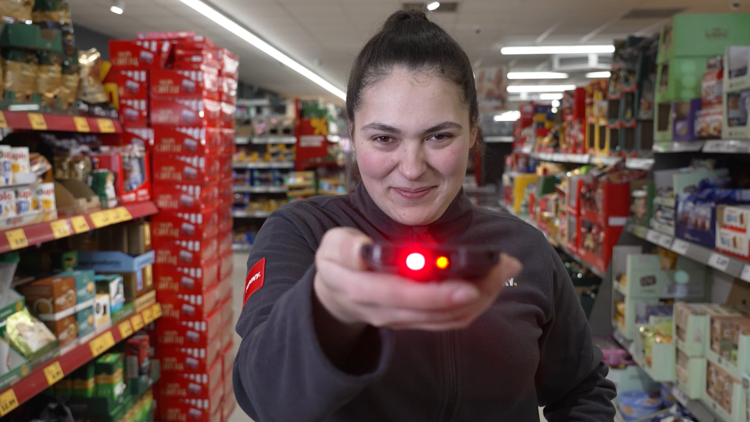 Penny Marktmitarbeiter:in die ein ZEBRA Gerät in die Kamera hält und zwischen Supermarktregalen steht