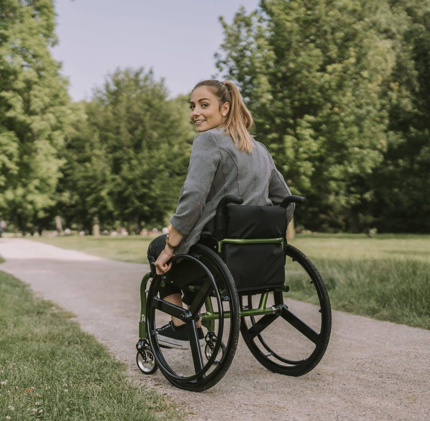 Junge Frau fährt im Rollstuhl durch den Park und lächelt in die Kamera.