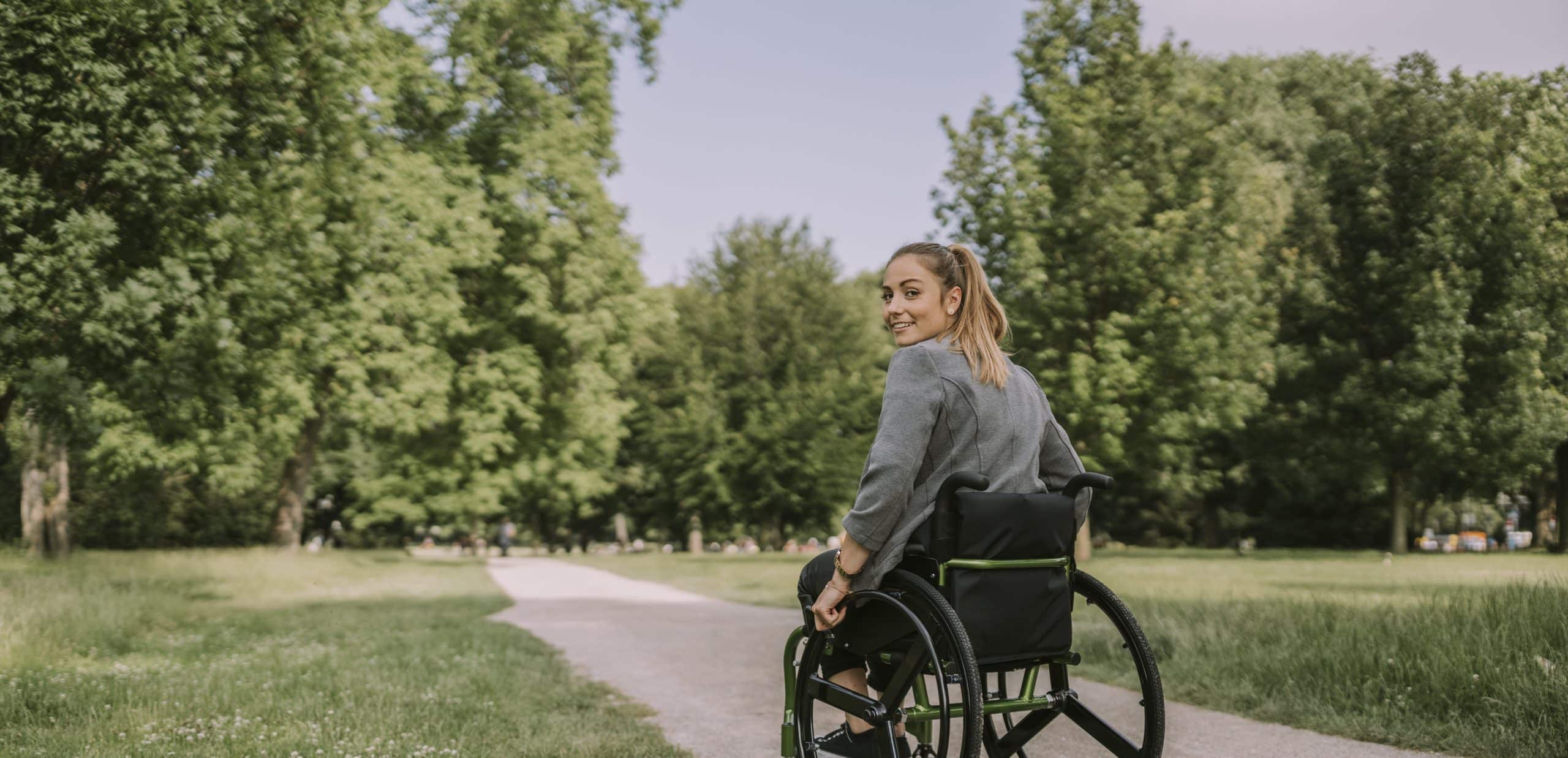 Junge Frau fährt im Rollstuhl durch den Park und lächelt in die Kamera.