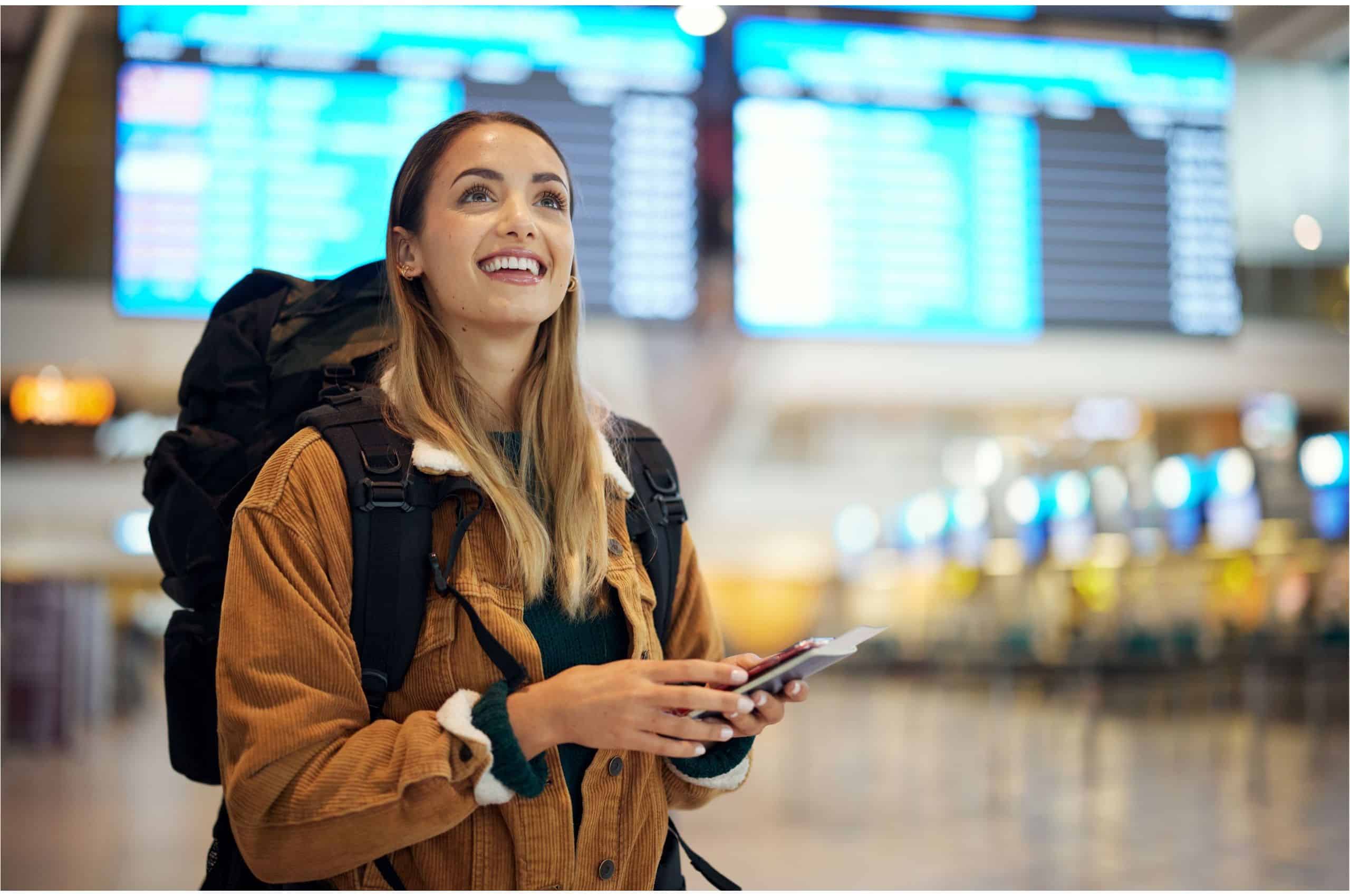 Junge Frau mit Rucksack hält Reisepass und Handy in der Hand. Im Hintergrund verschwommene Anzeigetafeln eines Flughafens.
