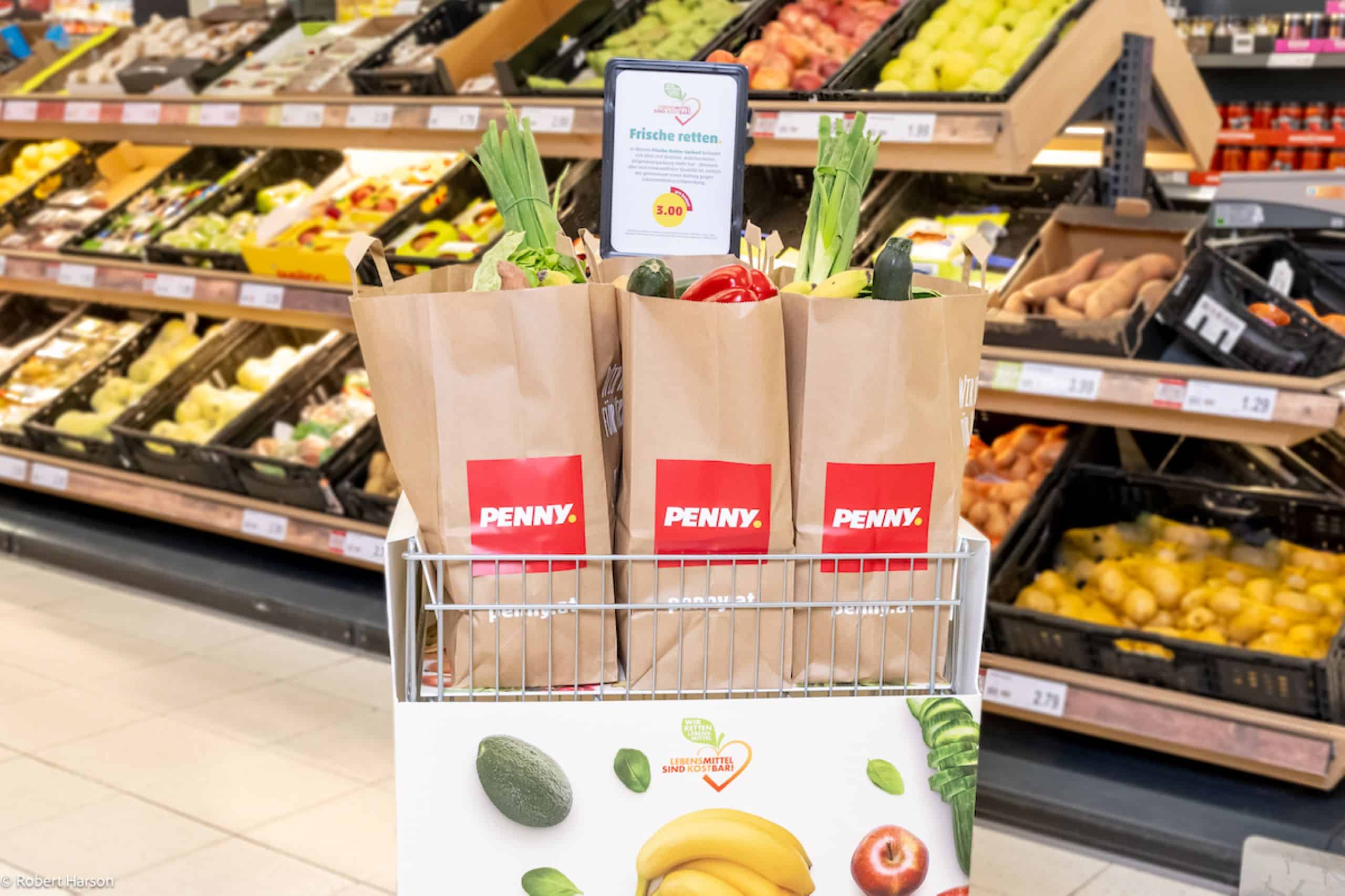 Papiertüten von PENNY gepackt mit Obst und Gemüse. Im Hintergrund die Gemüseabteilung eines PENNY Marktes.