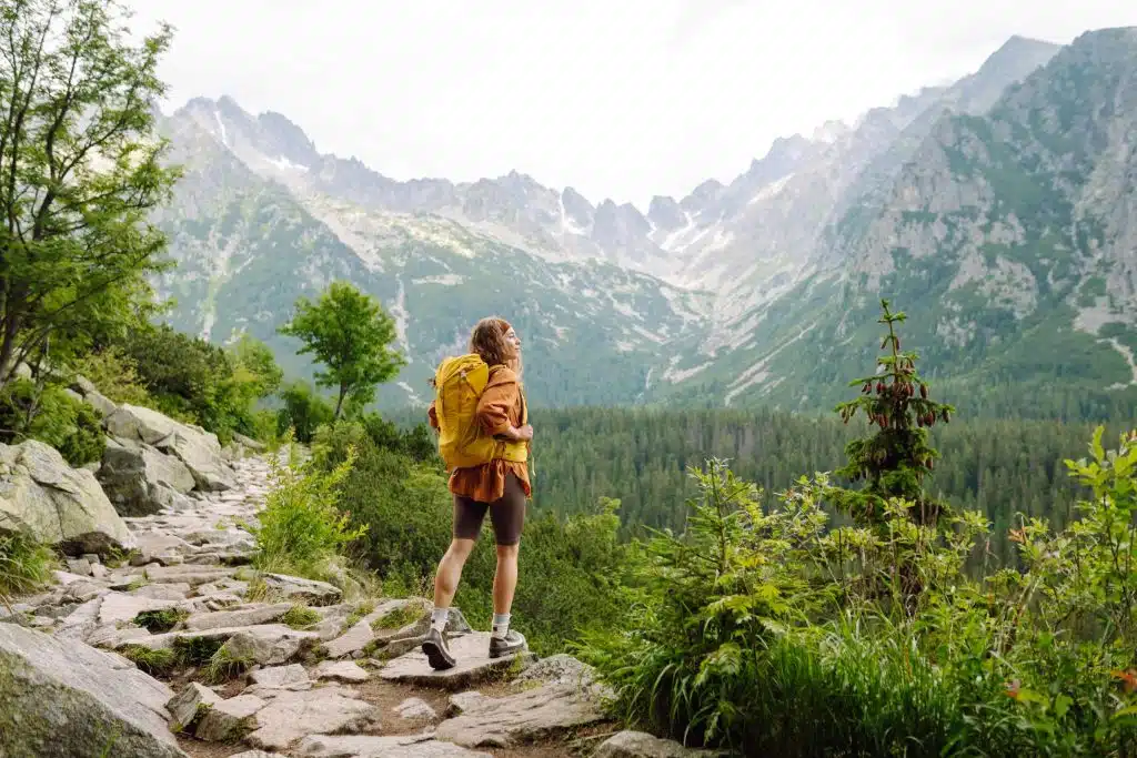 Frau mit einem gelben Wanderrucksack läuft über Wanderwege und blickt auf die Berglandschaft.