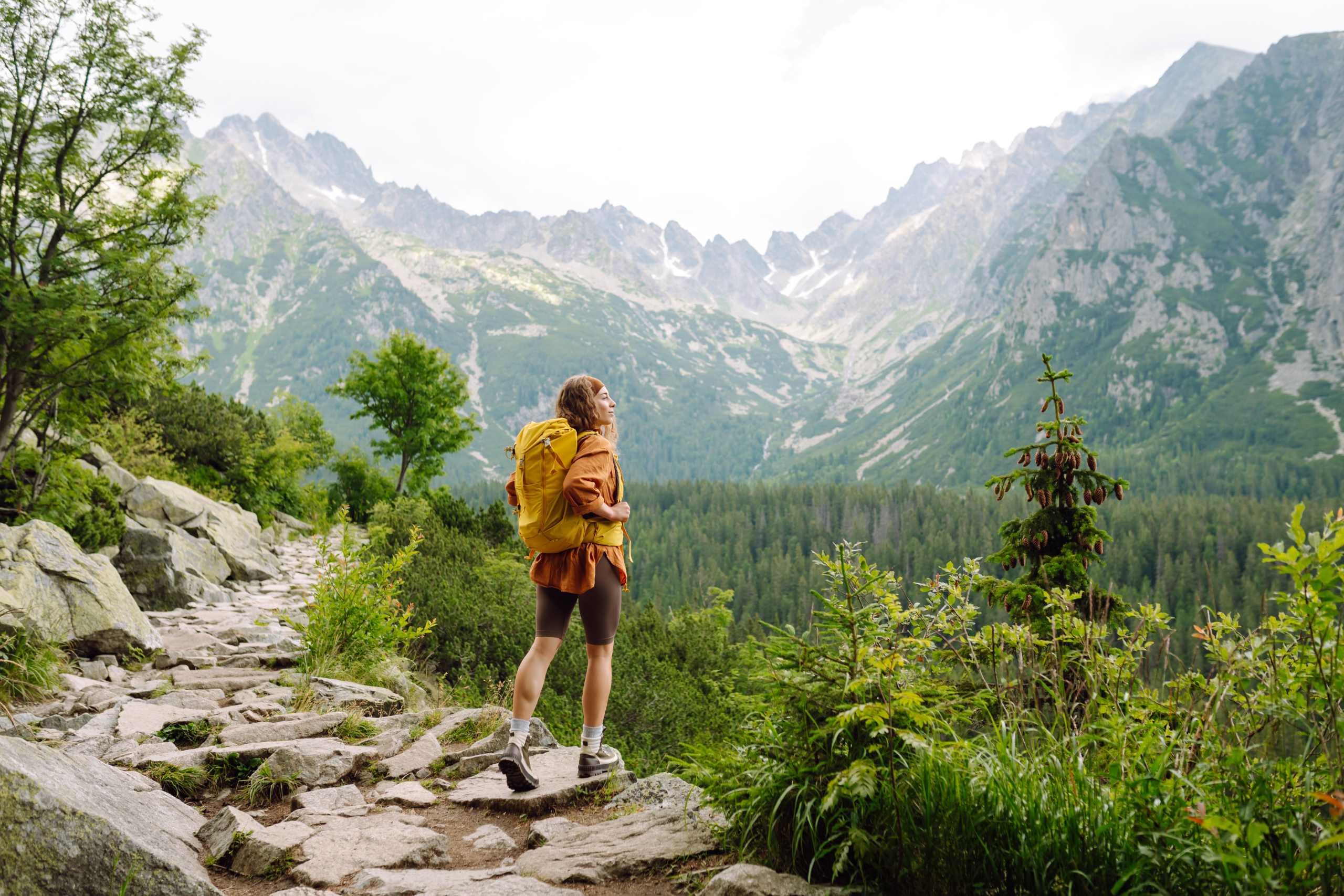 Frau mit einem gelben Wanderrucksack läuft über Wanderwege und blickt auf die Berglandschaft.