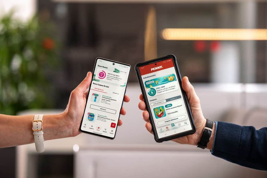 Zwei Hände halten Handys nebeneinander, auf denen jeweils die Apps von REWE und PENNY geöffnet sind