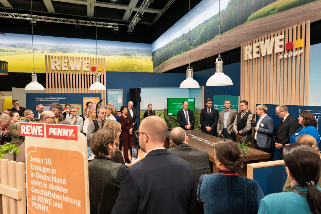 Seven people stand behind a desk at the REWE GROUP stand at the Green Week. One of the people is speaking to the audience via a microphone.