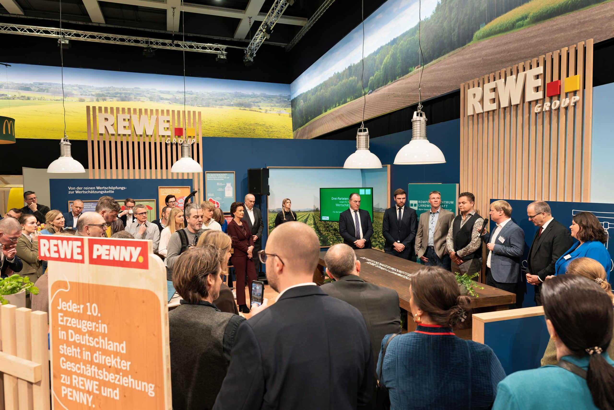 Seven people stand behind a desk at the REWE Group stand at the Green Week. One of the people is speaking to the audience via a microphone.