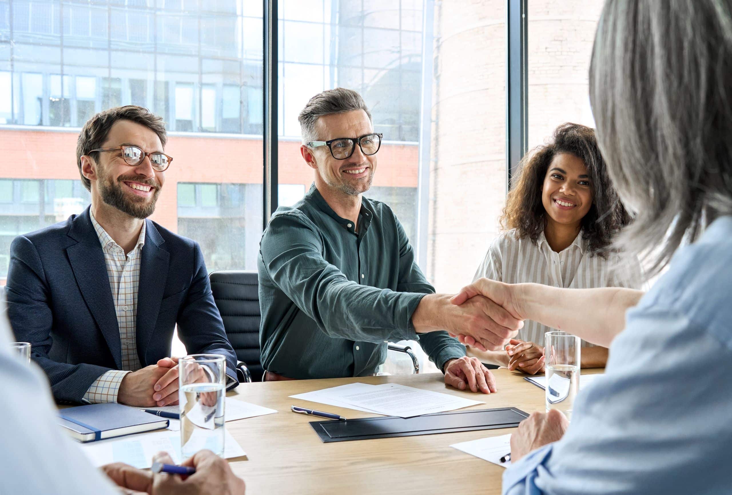 Four businesspeople are sitting at a table in an office, smiling warmly. Two of them are shaking hands.