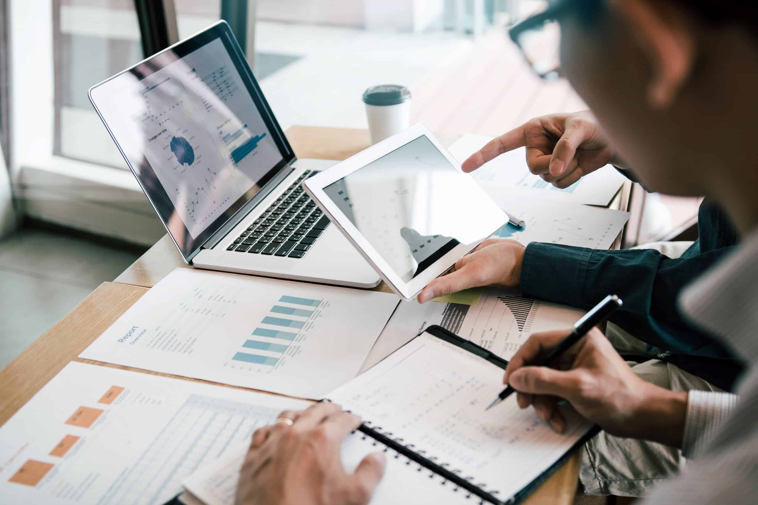 Two people have business documents and a laptop on the table in front of them and are looking at a tablet together.
