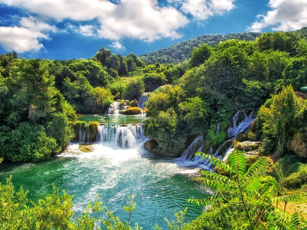 Mehrere Wasserfälle die in einem großen türkisenen See fließen. Um den See herum sind bewaldete Berge eines Nationalparks in Kroatien.