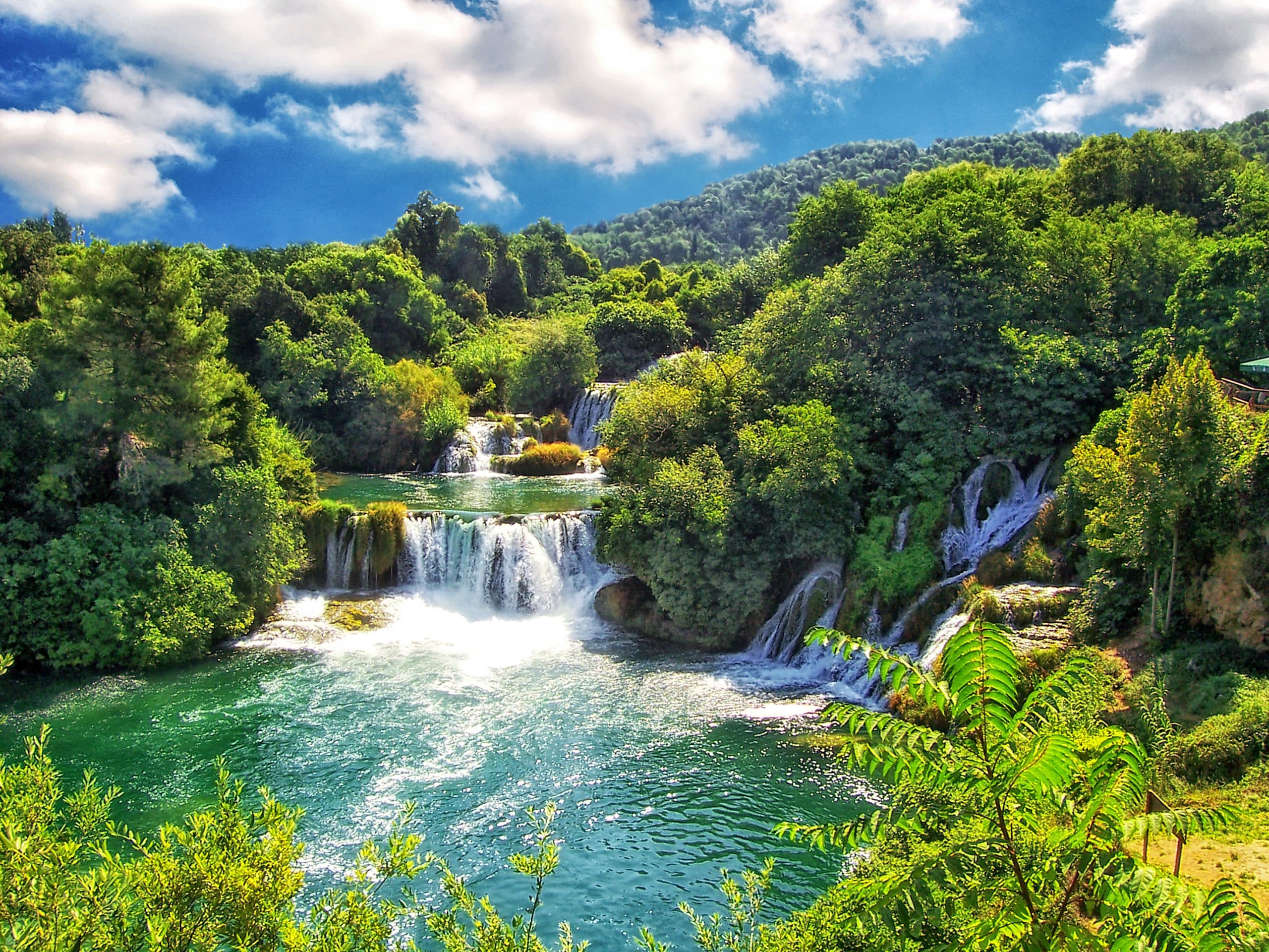Several waterfalls flowing into a large turquoise lake. Surrounding the lake are the forested mountains of a national park in Croatia.