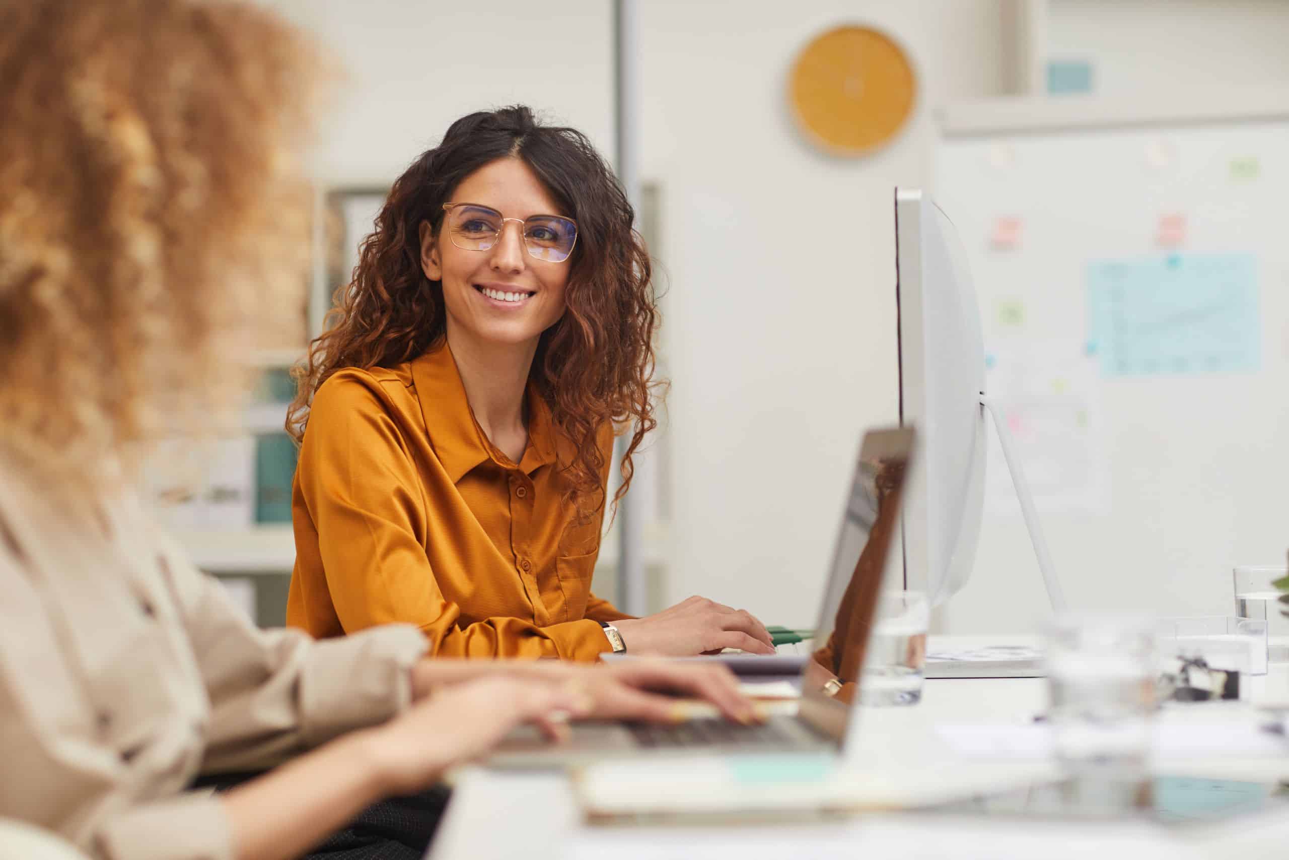 Zwei Frauen sitzen zusammen an ihren Computern im Büro. Eine Frau lächelt die andere freundlich an.