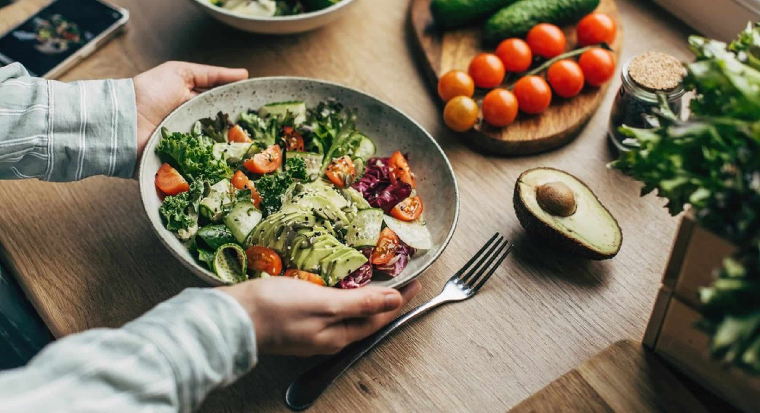 Eine Person hält einen Teller mit Salat in der Hand, auf dem Tisch liegen verschiedene Obst- & Gemüsesorten.