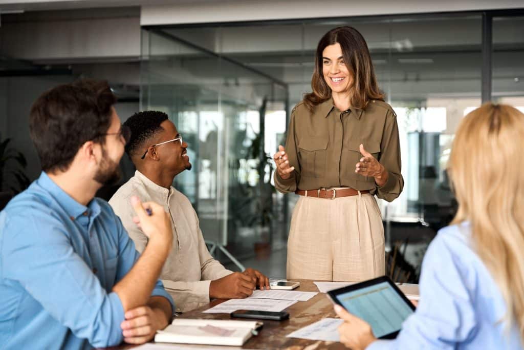 Eine Frau steht an einem Tisch im Büro und spricht lächelnd zu Mitarbeitenden am Tisch.