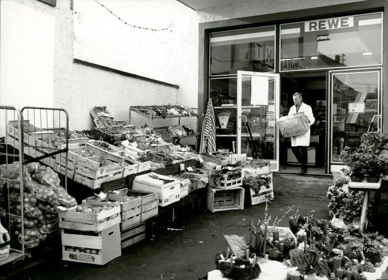 Ein schwarz-weiß Bild vom Eingangsbereich eines alten REWE Marktes. Ein Mitarbeiter im weißen Kittel trägt eine Kiste nach draußen, wo Obst und Gemüse in Holzkisten aufstellt sind.