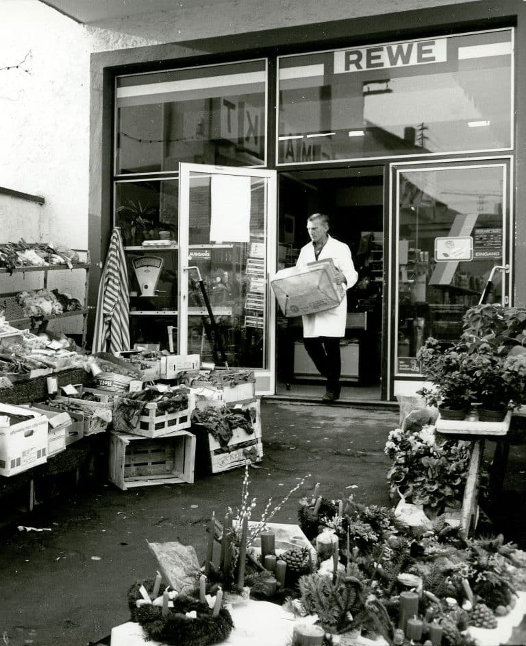 Ein schwarz-weiß Bild vom Eingangsbereich eines alten REWE Marktes. Ein Mitarbeiter im weißen Kittel trägt eine Kiste nach draußen, wo Obst und Gemüse in Holzkisten aufstellt sind.