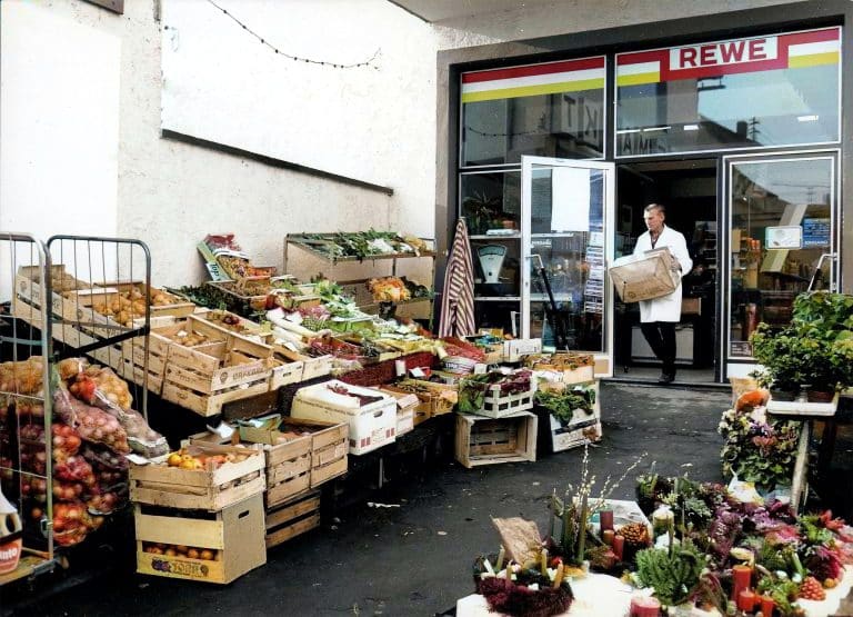 Ein nachkoloriertes Bild vom Eingangsbereich eines alten REWE Marktes. Ein Mitarbeiter im weißen Kittel trägt eine Kiste nach draußen, wo Obst und Gemüse in Holzkisten aufstellt sind.