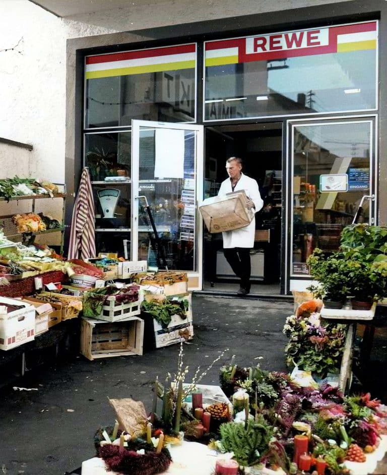 Ein nachkoloriertes Bild vom Eingangsbereich eines alten REWE Marktes. Ein Mitarbeiter im weißen Kittel trägt eine Kiste nach draußen, wo Obst und Gemüse in Holzkisten aufstellt sind.