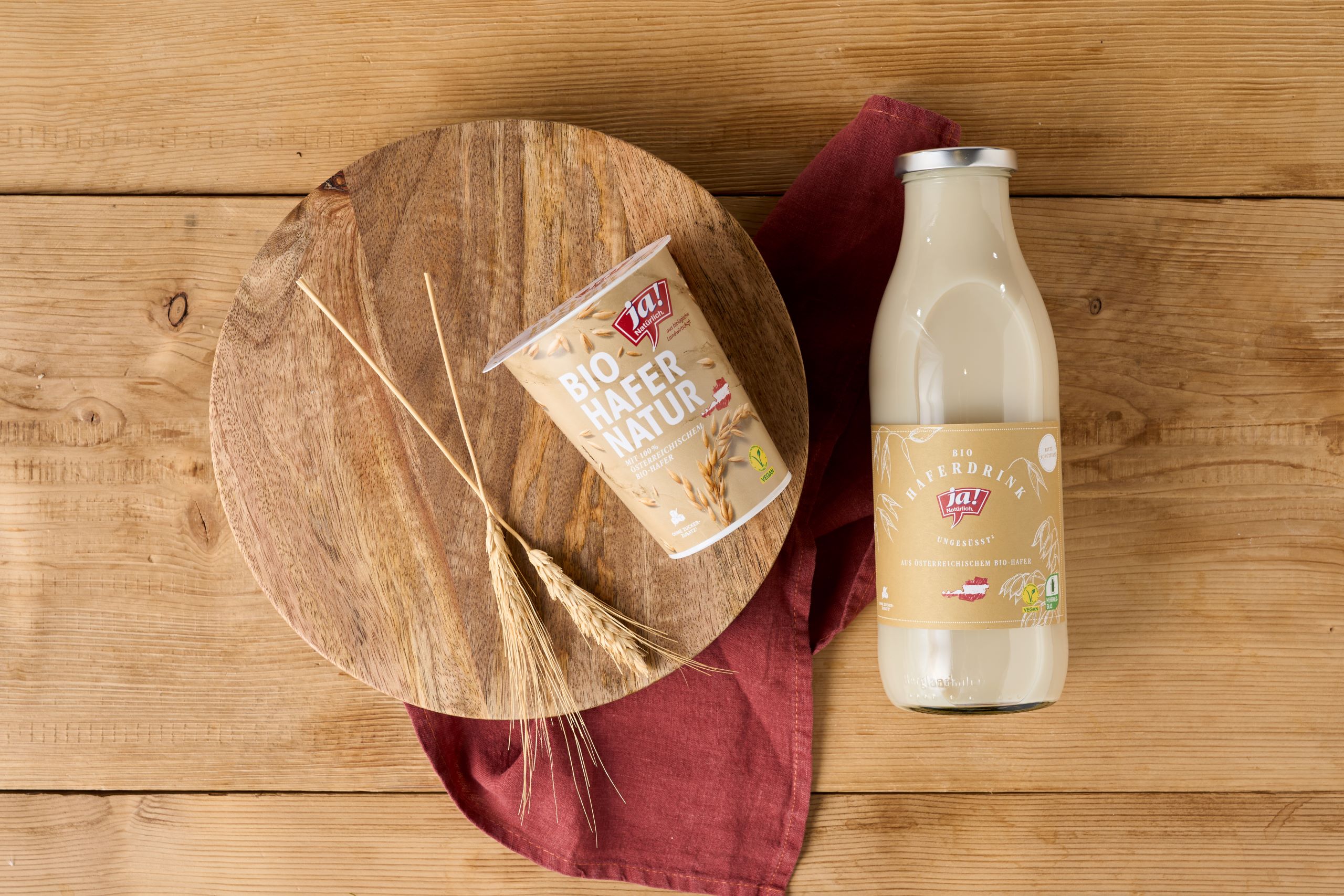 Ein Becher Bio-Haferjoghurt der Marke Ja! Natürlich liegt auf einer Holzplatte mit Hafer als Deko. Daneben liegt eine Glasflasche mit Bio-Haferdrink.