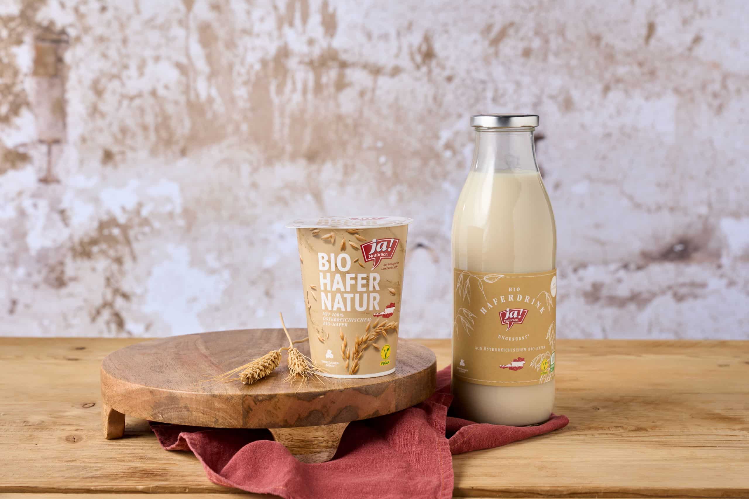 Ein Becher Bio-Haferjoghurt der Marke Ja! Natürlich steht auf einer Holzplatte mit Hafer als Deko. Daneben steht eine Glasflasche mit Bio-Haferdrink.