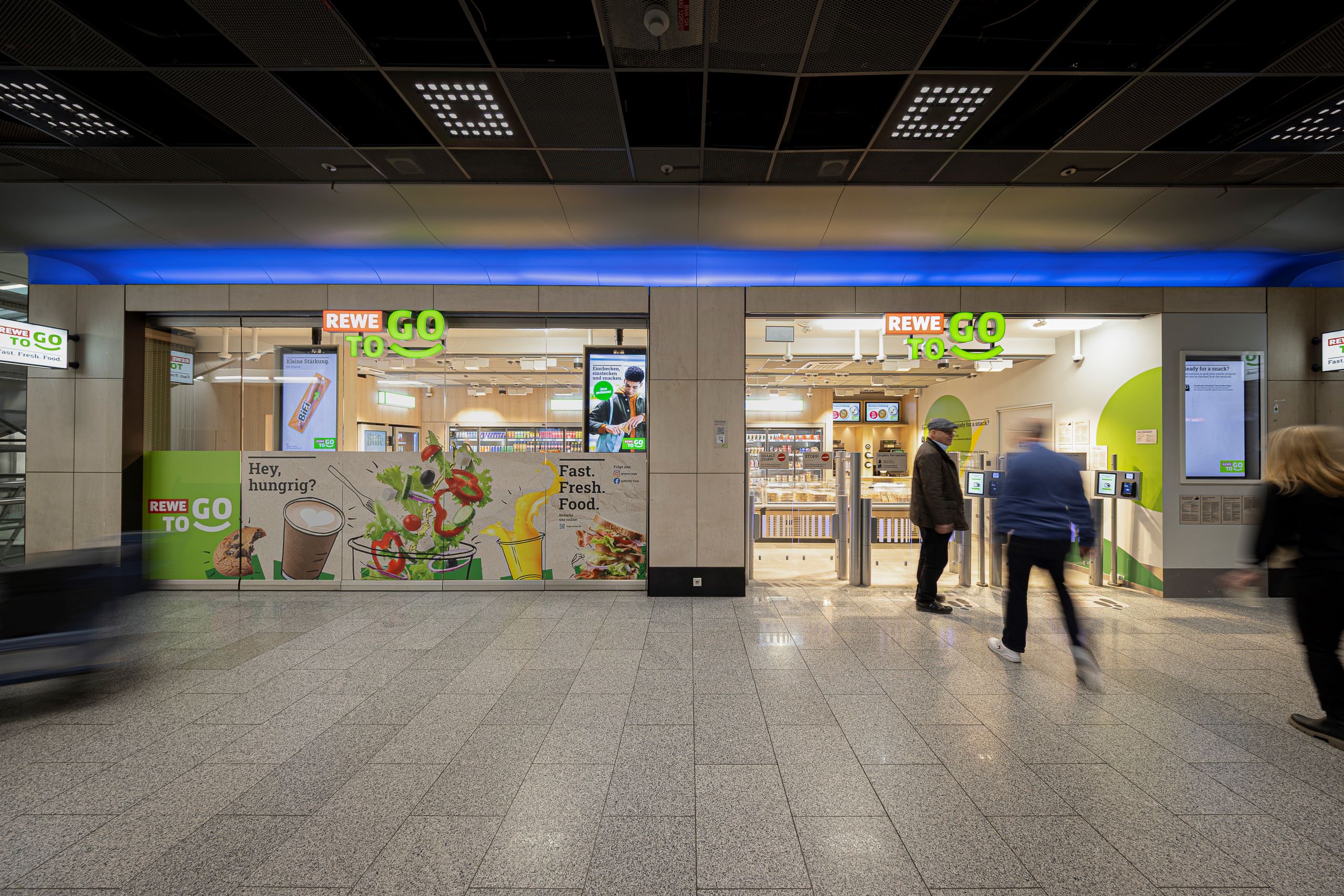 Außenansicht des REWE To Go am Flughafen Frankfurt. 3 Personen sind verschwommen vor dem Markt zu sehen.