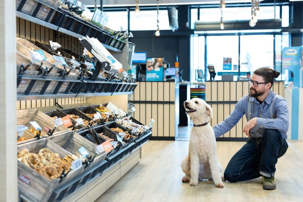 Ein Mann kniet neben seinem Hund in einem ZooRoyal Markt an der Snackbar, die unverpackte Leckereien für Haustiere bietet.
