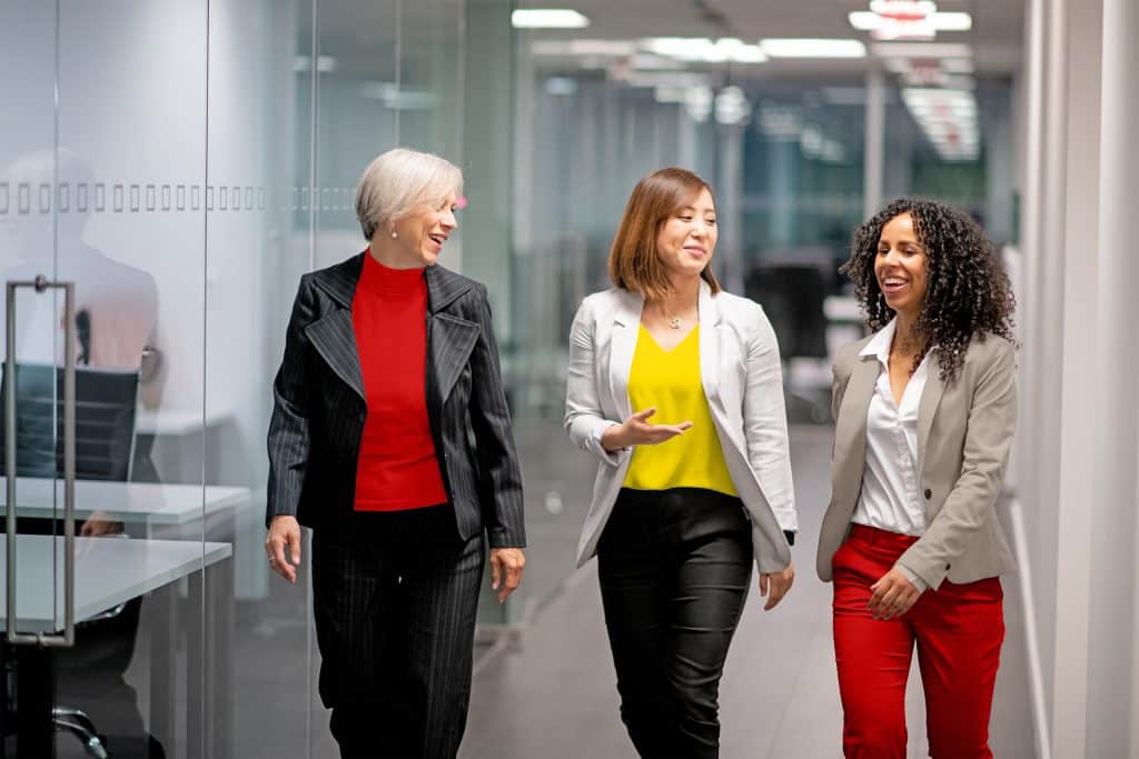 Drei Frauen laufen durch einen Flur im Büro und unterhalten sich lächelnd.