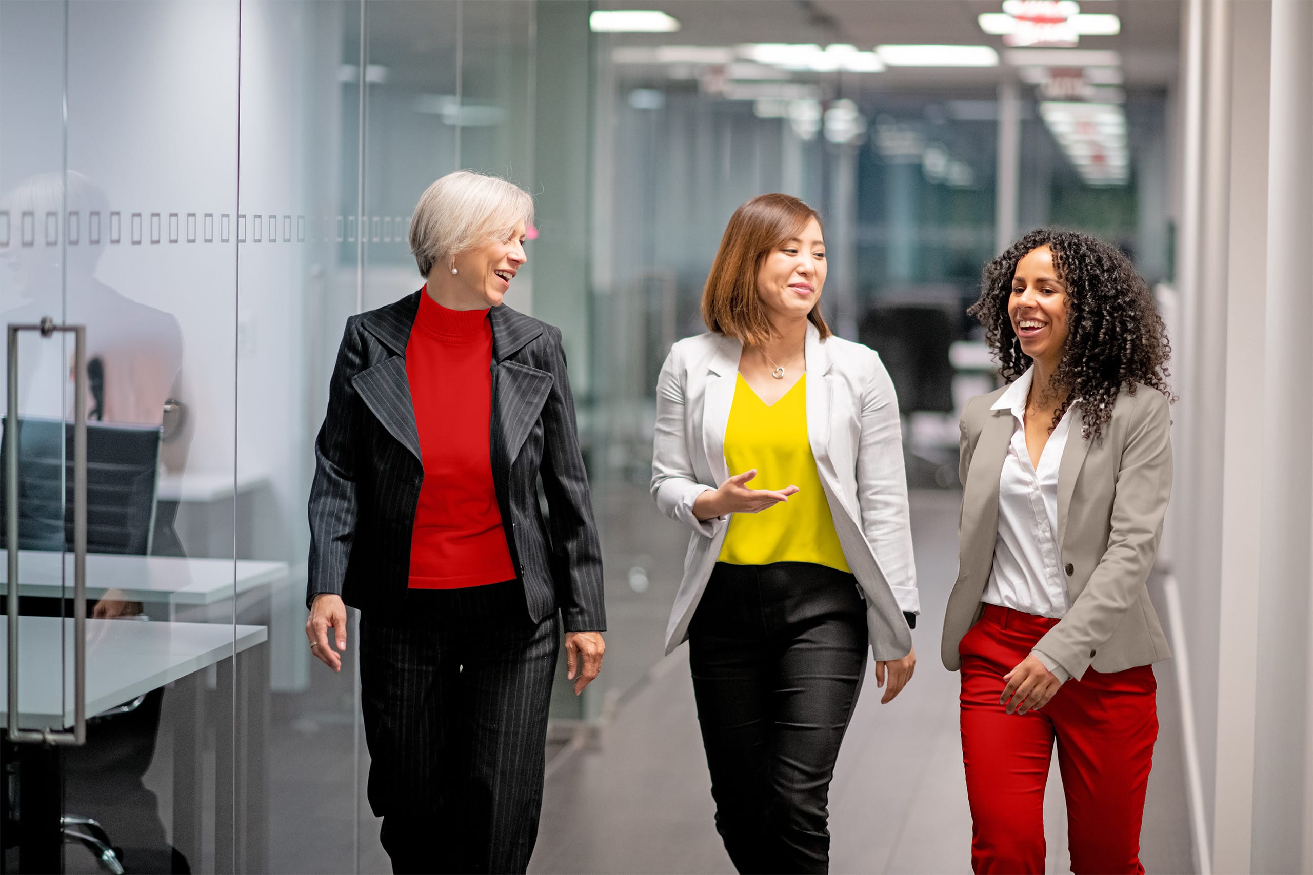 Drei Frauen laufen durch einen Flur im Büro und unterhalten sich lächelnd.