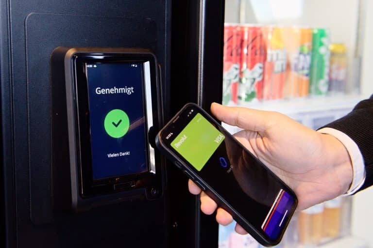 Eine Person hält ein Handy zum kontaktlosen bezahlen an das Lesegerät von einem Getränkeautomaten.