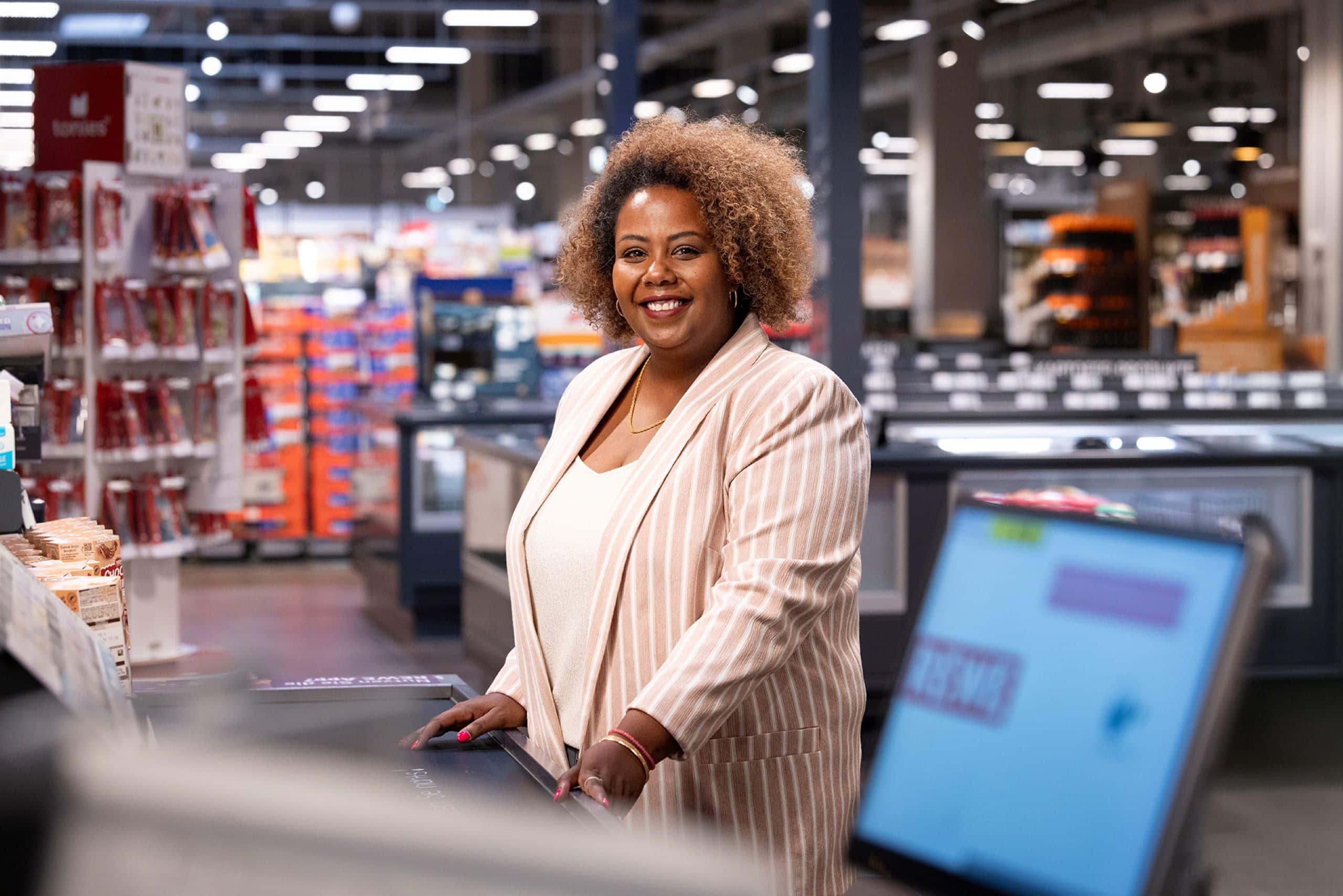 Porträt von Semai Akale. Sie steht in ihrem REWE Markt an der Kasse und lächelt in die Kamera.