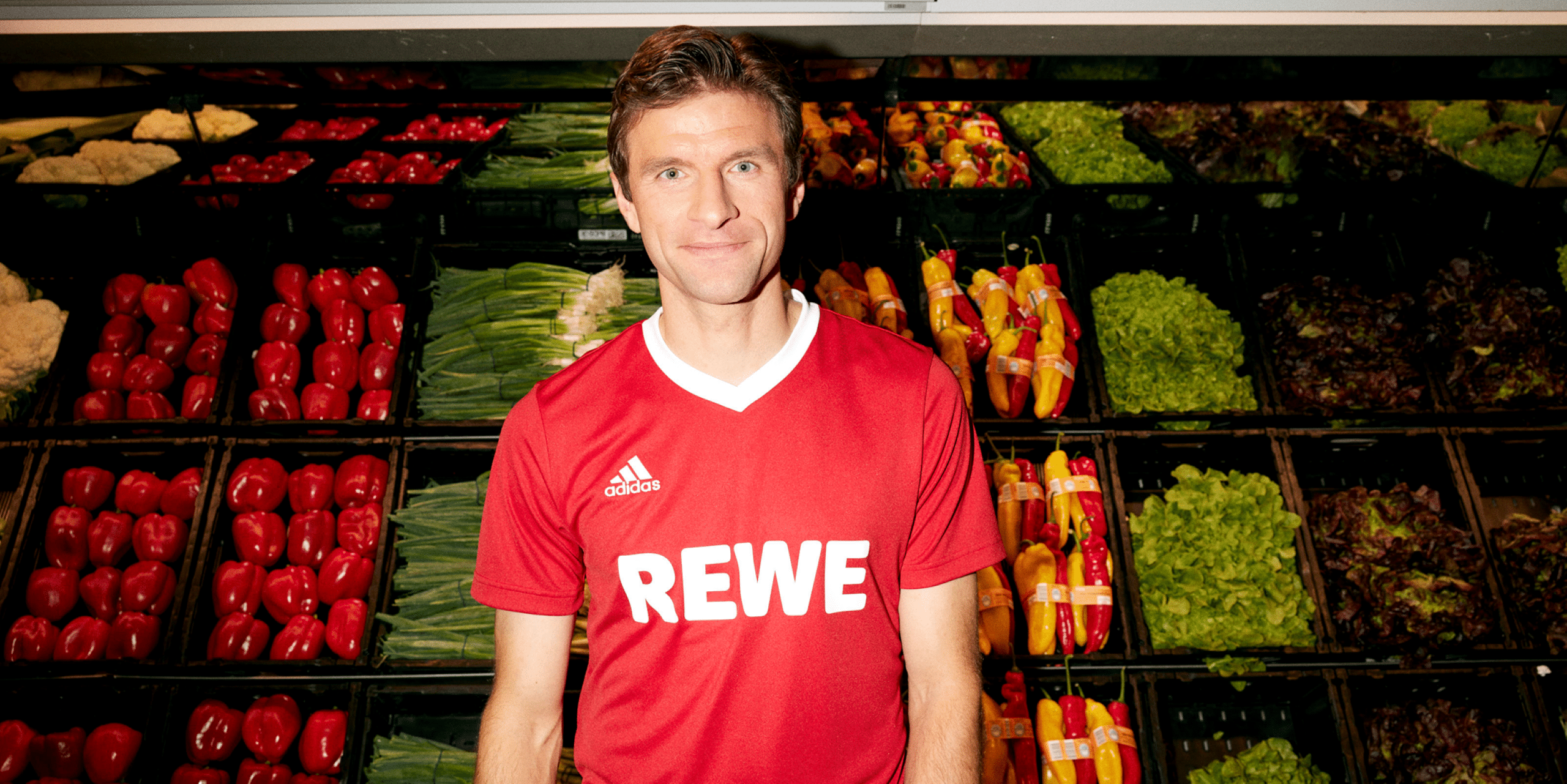 Porträt von Thomas Müller. Er trägt ein rotes Trikot mit dem REWE Logo und steht in der Gemüseabteilung eines REWE Marktes.