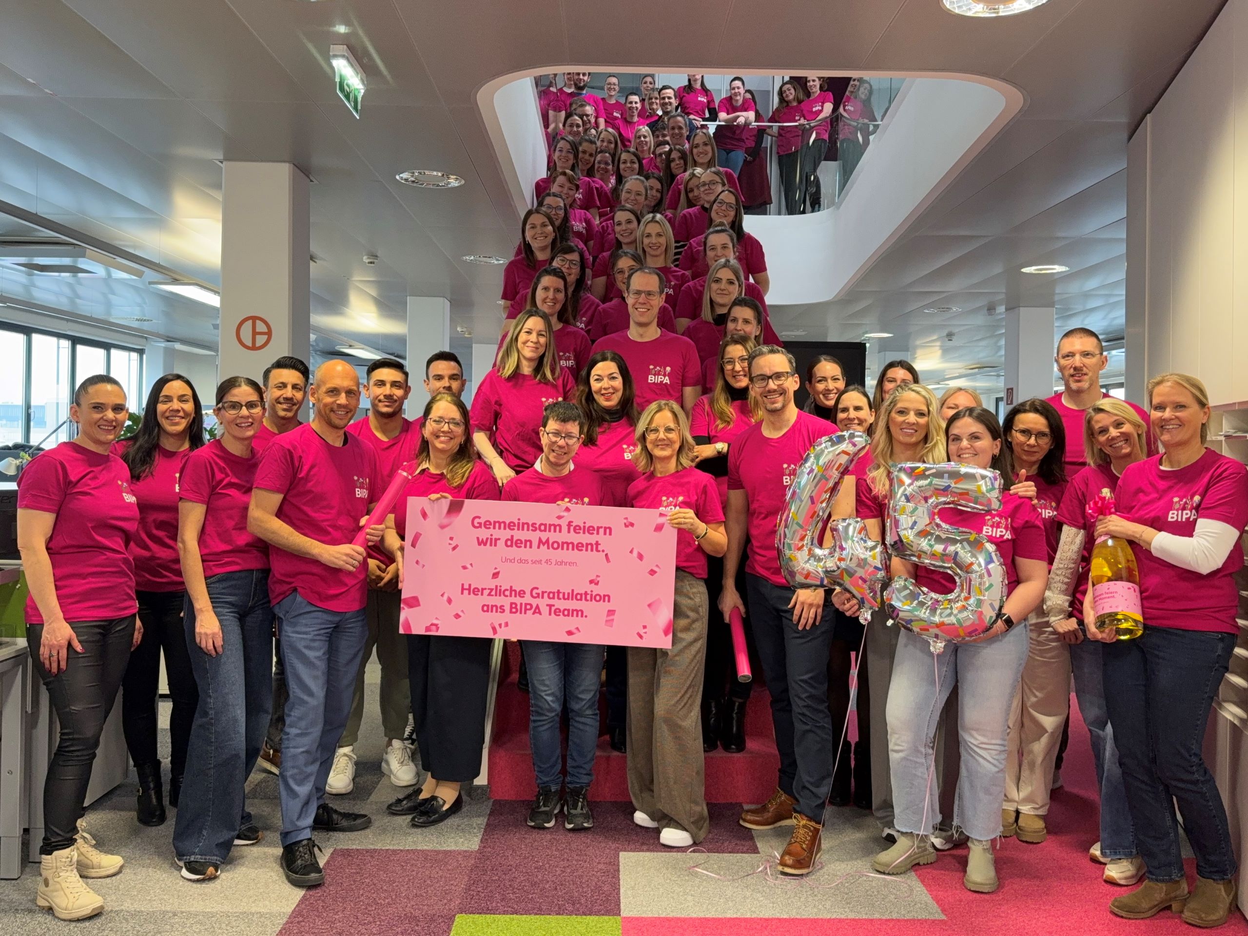 Eine große Gruppe von BIPA Mitarbeiter:innen, die alle pinke T-Shirts mit dem BIPA-Logo tragen, steht an einer Treppe aufgestellt. In der ersten Reihe halten Mitarbeiter:innen Ballons und ein Schild zur Feier Feier des 45. Jubiläums.