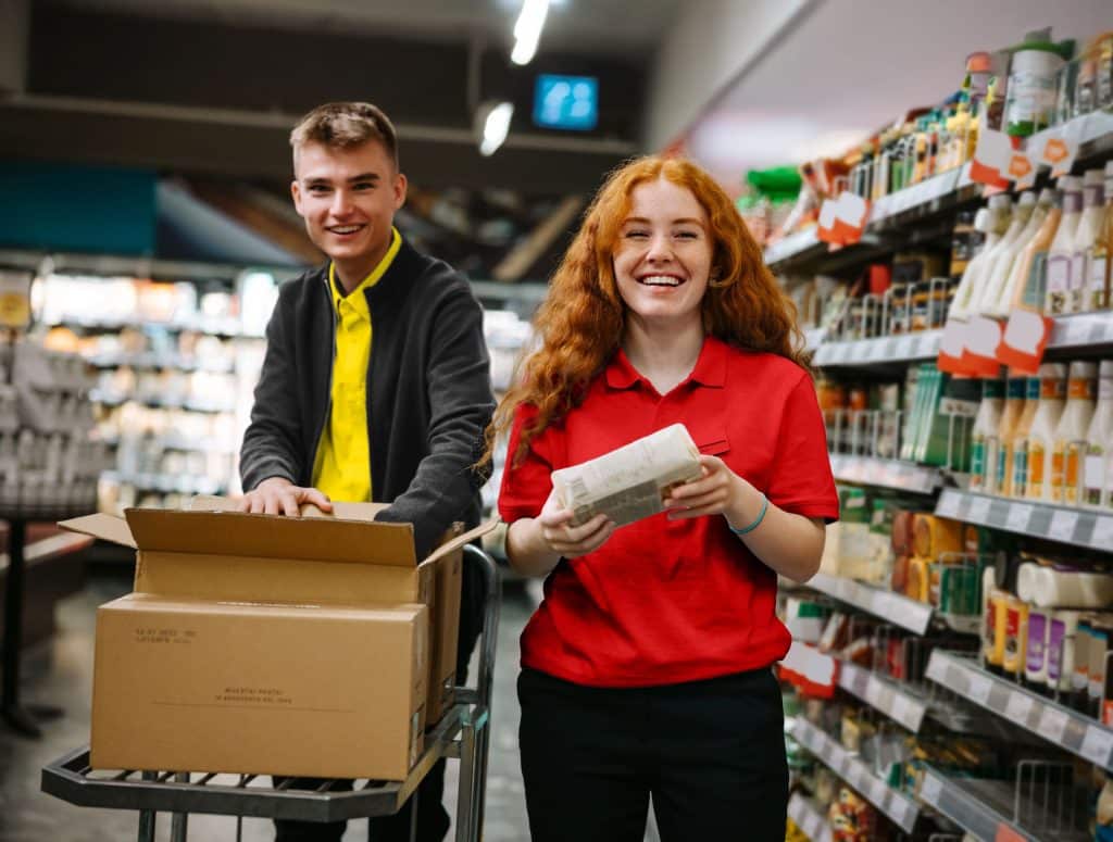 Zwei Mitarbeiter:innen eines Supermarktes bei der Arbeit. Eine junge Frau in einem roten Shirt und ein junger Mann in einem gelben Shirt stehen lachend nebeneinander und räumen Ware ein.
