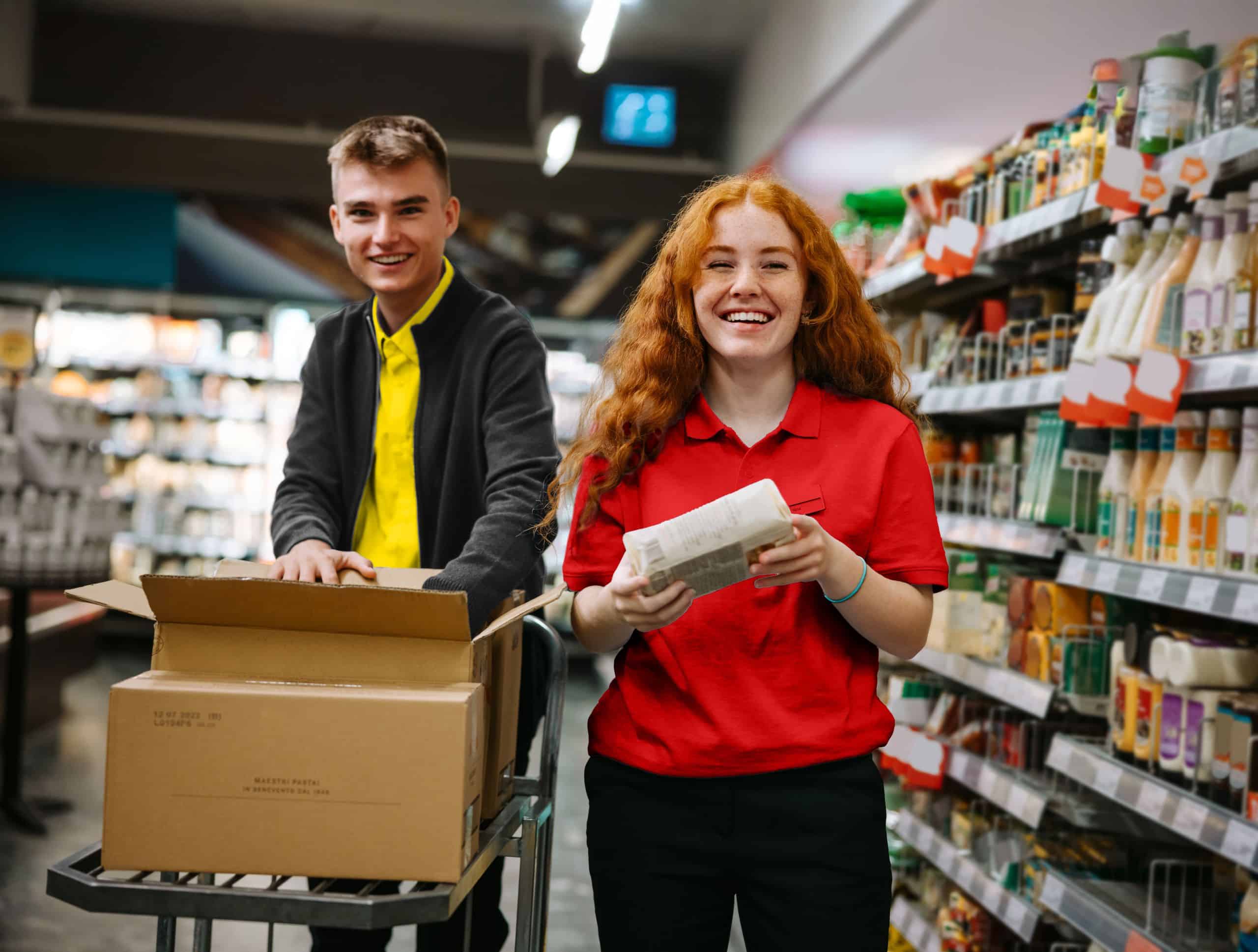 Zwei Mitarbeiter:innen eines Supermarktes bei der Arbeit. Eine junge Frau in einem roten Shirt und ein junger Mann in einem gelben Shirt stehen lachend nebeneinander und räumen Ware ein.