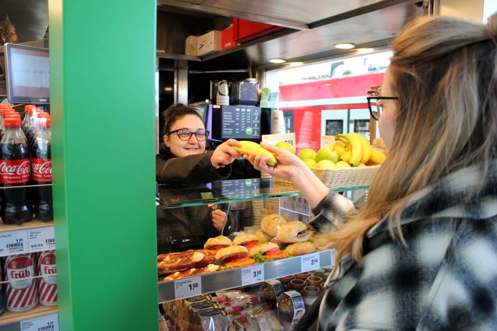 Eine Frau kauft am REWE To Go Gleiskiosk ein und bekommt von der Verkäuferin eine Banane gereicht.