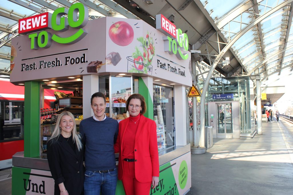 Helen Schnürle, Michael Mayer-Sonnenburg and Jenny Ramme stand in front of a REWE To Go kiosk on the platform at Cologne Central Station and smile into the camera.