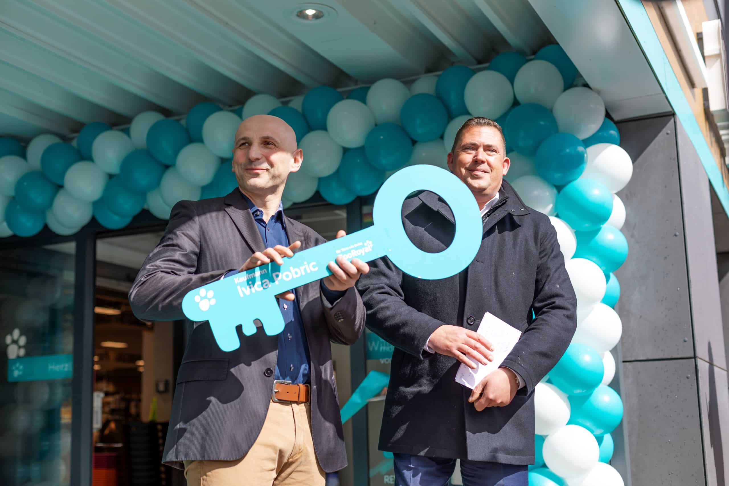 ZooRoyal Kaufmann Ivica Pobric hält einen großen blauen Schlüssel aus Pappe in den Händen, anlässlich der Eröffnung seines ZooRoyal Marktes, der mit Luftballons geschmückt im Hintergrund zu sehen ist. Neben ihm steht Jochen Vogel, Vorsitzender der Geschäftsleitung Region Nord.