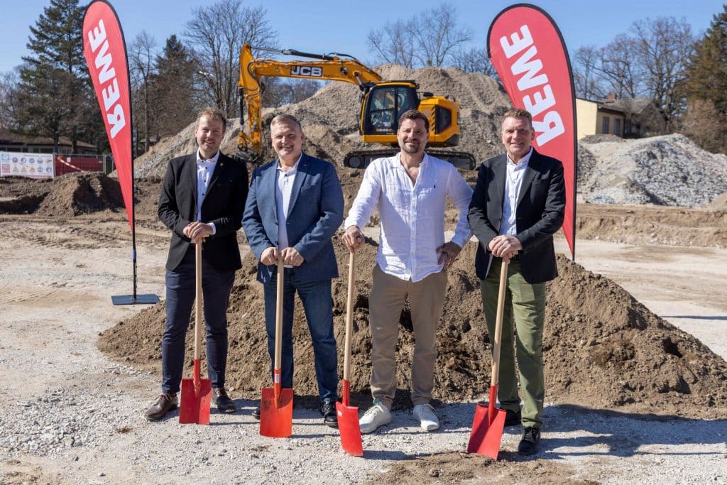 Vier Männer stehen mit Schaufeln in der Hand auf einer Baustelle und lächeln in die Kamera. Im Hintergrund ist ein Bagger zu sehen und zwei REWE Flaggen.