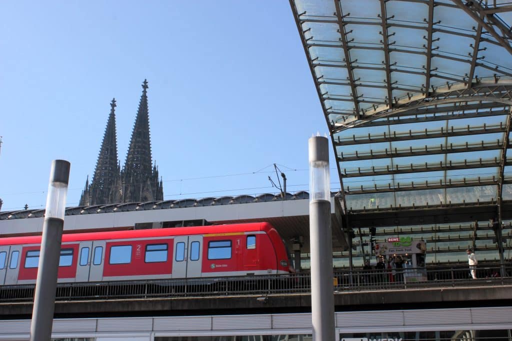 Bild von unten mit Blick nach oben auf den REWE To Go Gleiskiosk am Kökner Hauptbahnhof mit dem Kölner Dom im Hintergrund.